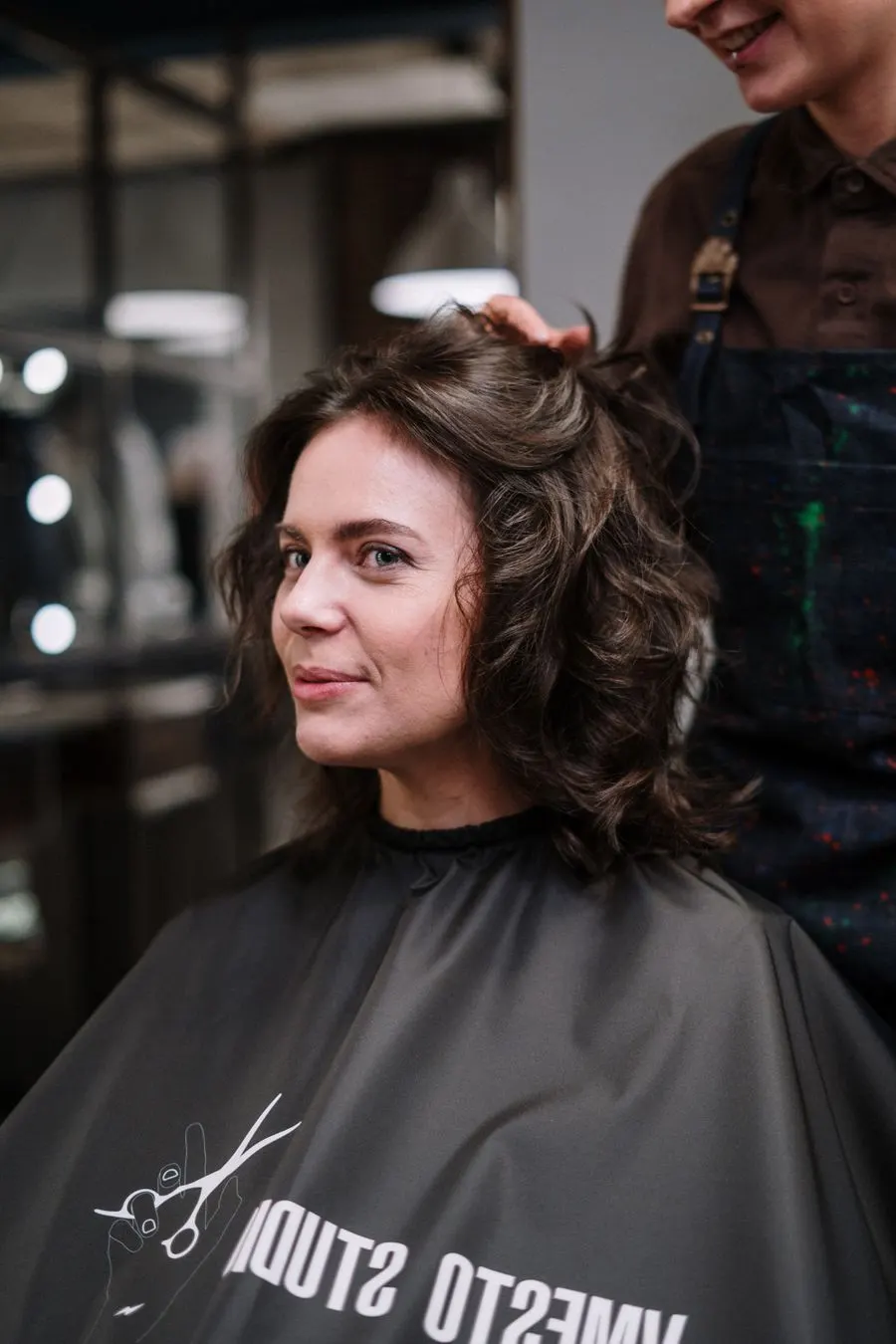 Cliente souriante aux cheveux bruns bouclés après sa coupe, la coiffeuse derrière elle