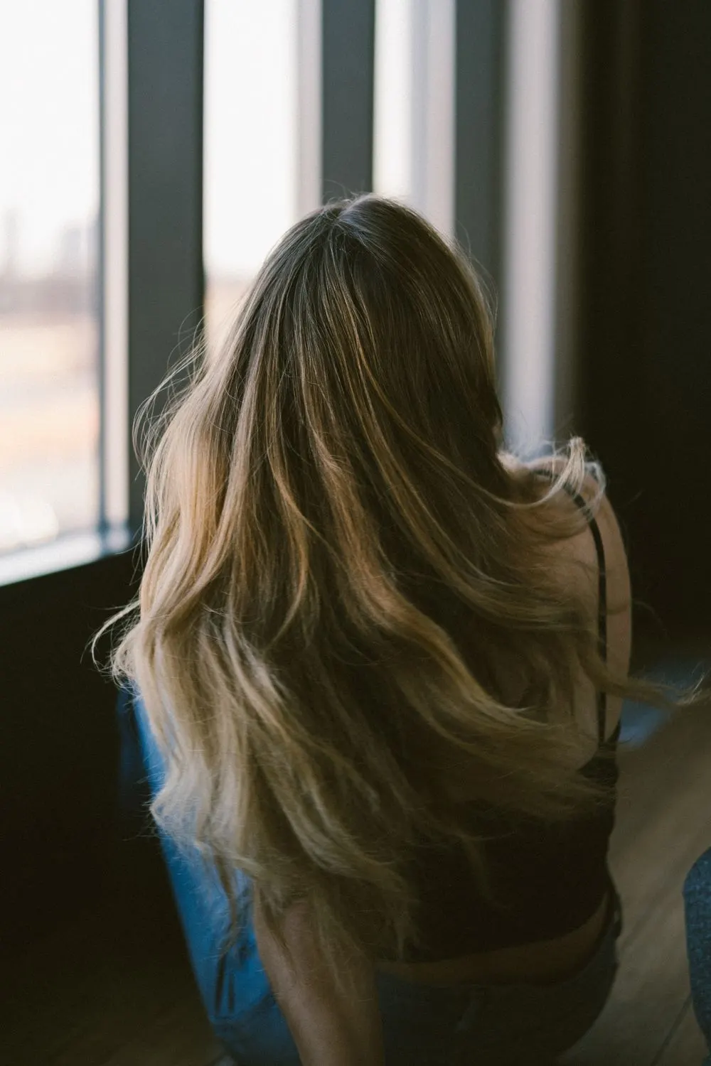 Longs cheveux ondulés éclaircis, vue de dos près d’une fenêtre
