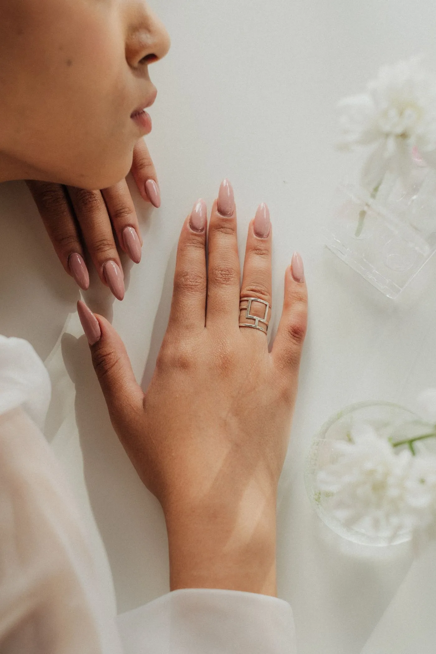 Main posée sur une surface blanche, ongles amande nude brillants, bague argent géométrique, lumière douce
