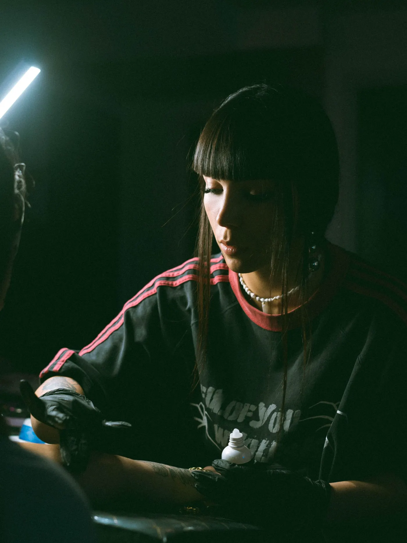 La tatoueuse Lou au travail, frange noire, gants noirs, sous une lampe froide dans le studio sombre
