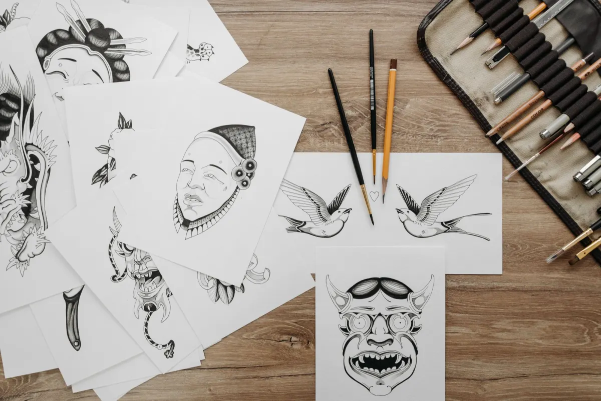 Dessins de tatouage à l’encre étalés sur une table en bois, hirondelles, masque, visage, pinceaux