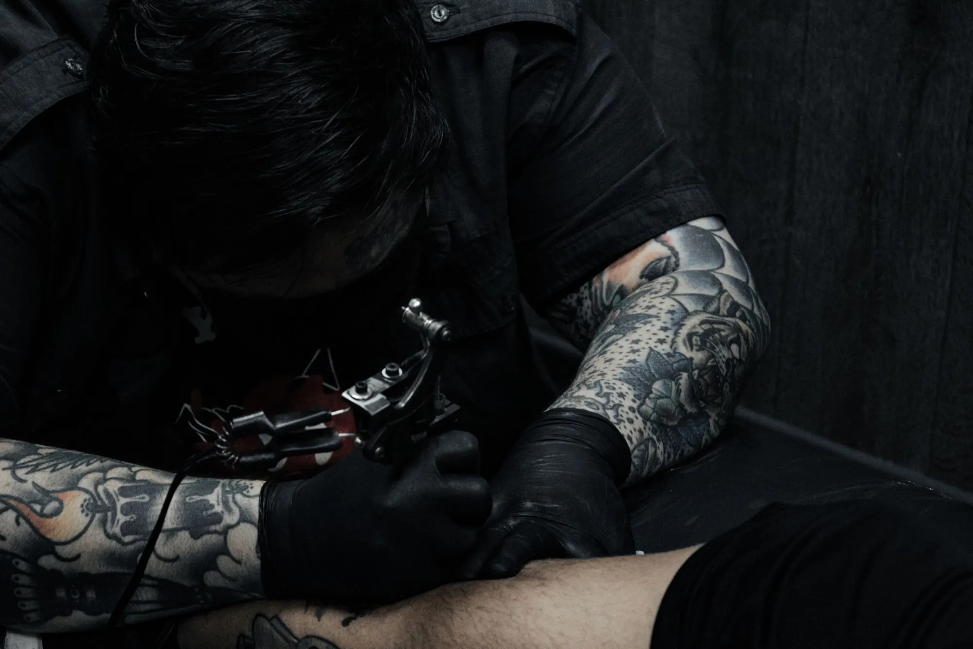 Tatoueur penché sur l’avant-bras d’un client, gants noirs, machine à bobines, bras couverts de tatouages, lumière de studio sombre