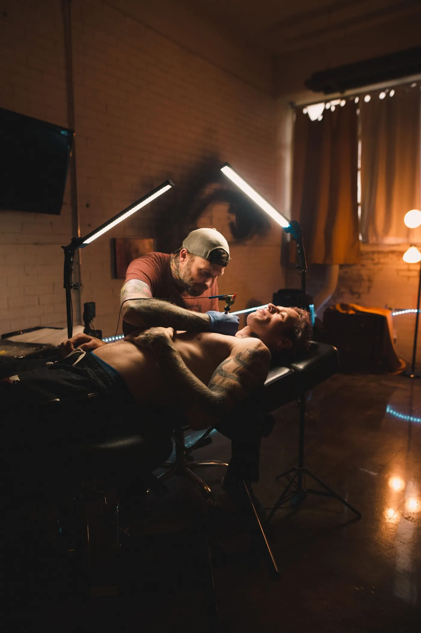 Salle de tatouage dans la pénombre, deux lampes allumées au-dessus d’une séance