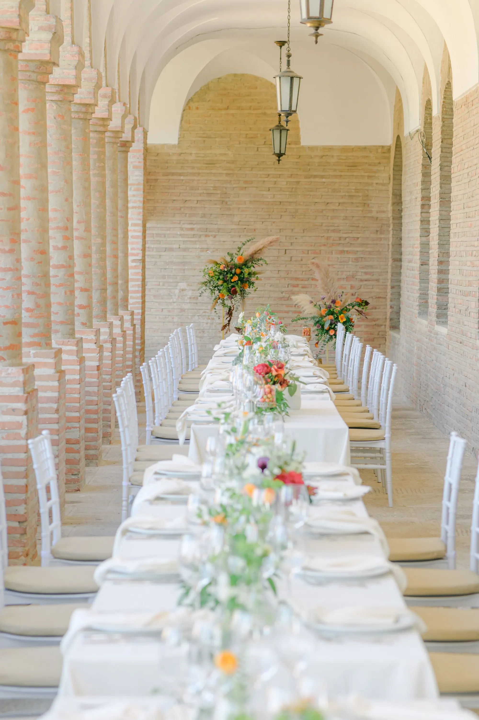 Longue table de réception dressée sous une galerie, nappes claires, verres en cristal et fleurs blanches